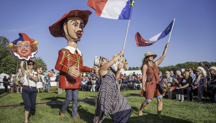 A group of people outdoors in a field march holding homemade large traditional Acadian celebratory masks and a large Acadian flag