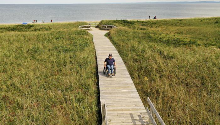 a person using a wheelchair appears on a baordwalk approaching a beach in Nova Scotia with beach grass extending along both sides of the long wooden boardwalk